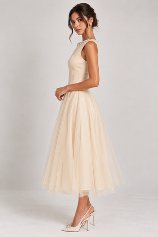 A woman models a sleeveless champagne tulle midi dress with a high neckline and a full, flowing skirt, paired with white slingback heels against a light gray studio background.