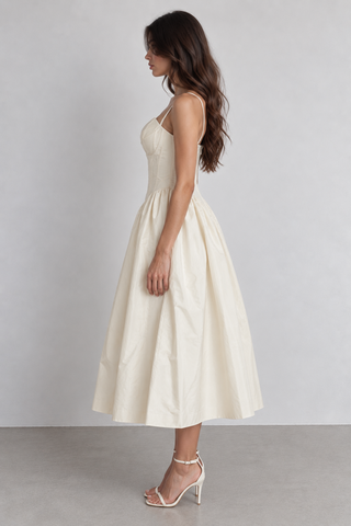 A woman modeling a cream-colored midi dress with spaghetti straps and a flared skirt, standing in profile against a light gray studio background.