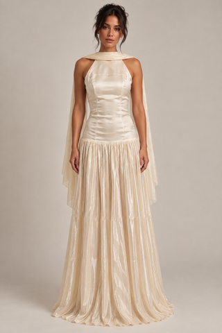 A model wearing a floor-length cream satin gown with a halter neckline, draped cape sleeves, and a pleated tiered skirt, photographed against a neutral beige studio background.