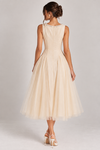 A woman modeling a sleeveless cream-colored midi dress with a structured waist and a full tulle skirt, shown from the back against a light gray studio background.