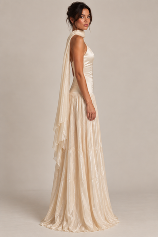 A woman modeling a sleeveless champagne-colored maxi dress with a high neck, pleated skirt, and a draped cascading overlay, shown against a neutral studio backdrop.