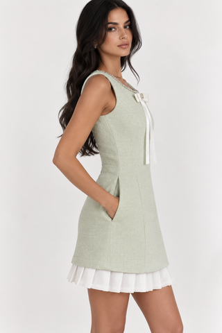 A woman modeling a sleeveless light sage green dress with a white ruffled hem and a white bow at the neckline, photographed against a plain light gray studio background.