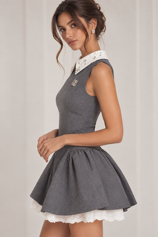 A woman modeling a sleeveless gray mini dress with a white lace collar and a flared pleated skirt with a white lace hem, photographed against a soft light gray studio backdrop.