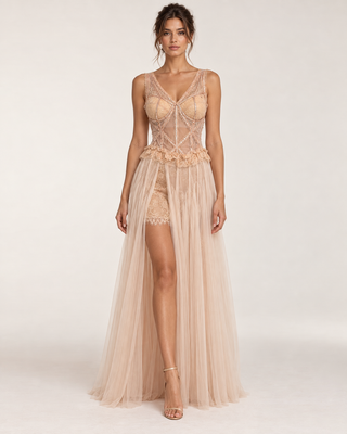 A model wears a champagne beige lace and tulle evening gown with a sheer corset bodice, floral waist detail, and a long flowing tulle overskirt, photographed against a plain light background.