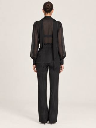 A model shown from the back wearing a black sheer long-sleeve top with a structured collar and draped front, paired with black pinstripe trousers, against a light gray studio background.