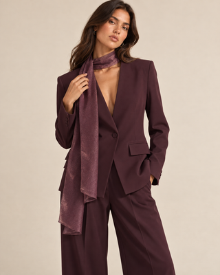 A woman models a matching deep plum purple tailored blazer and wide-leg trousers set, styled with a matching textured scarf against a plain beige studio background.