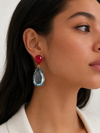 A close-up of a woman wearing a dangling earring with a faceted red gemstone and a large teardrop-shaped light blue gemstone in a gold setting.