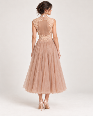 A woman shown from the back wearing a sleeveless midi dress with a sheer lace back bodice and a full tulle skirt in a dusty blush pink color, paired with nude heels, against a light gray studio background.