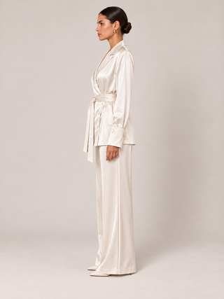 A woman stands in profile wearing a matching cream satin wrap jacket and wide-leg trousers set against a plain light beige studio background.