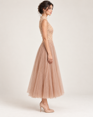 A woman modeling a sleeveless, high-neck midi dress with a lace bodice and a voluminous tulle skirt in a dusty blush pink color, paired with clear high-heeled shoes, against a light beige studio background.