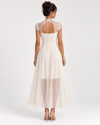 A woman viewed from the back wearing a sleeveless lace dress with a high open back, cap sleeves, and a flowing tulle skirt, photographed against a plain light gray studio background.