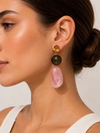 A close-up of a woman wearing a dangling statement earring with a gold-toned top, a dark green round stone, and a large pink teardrop stone drop.