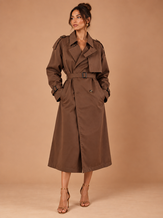A woman modeling a mid-length brown trench coat with a matching belt, photographed against a solid warm beige studio backdrop.
