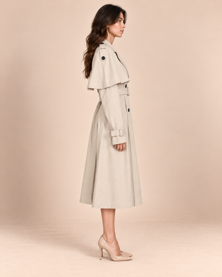 A woman modeling a light beige trench coat with a matching belt and long sleeves, shown in profile against a solid beige studio background.