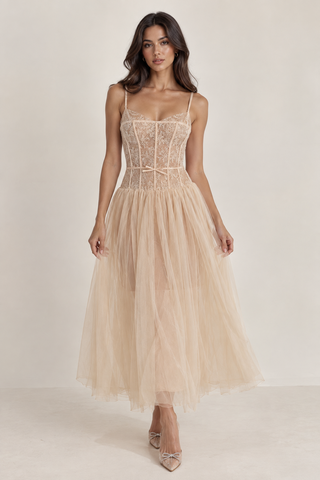 A woman modeling a sleeveless midi dress with a structured lace corset bodice and a voluminous tulle skirt in a light champagne-beige color, set against a plain cream studio background.