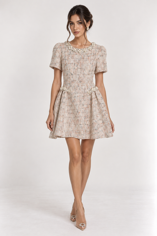 A woman models a short-sleeve tweed mini dress with a pleated skirt and pearl embellishments on the neckline and waist, set against a plain light gray studio background.