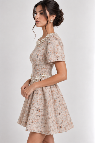 A woman modeling a short-sleeve tweed dress with pearl embellishments at the neckline and waist, shown against a plain light gray studio background.