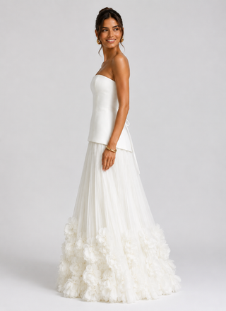 A woman modeling a strapless white wedding gown with a structured bodice, sheer overlay, and a voluminous ruffled tulle skirt against a light gray studio background.