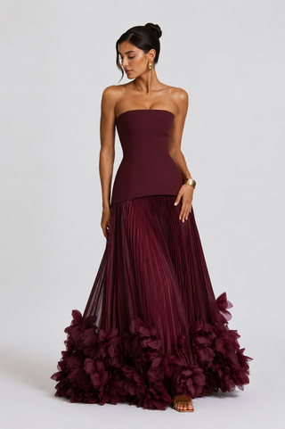 A woman models a strapless floor-length gown in deep burgundy. The bodice is structured and fitted, while the full skirt is voluminous and pleated, finished with a dramatic ruffled floral hem.
