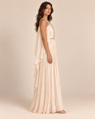 A woman modeling a sleeveless, floor-length pleated gown in a soft champagne beige color, shown from a side-back angle against a plain beige studio backdrop.