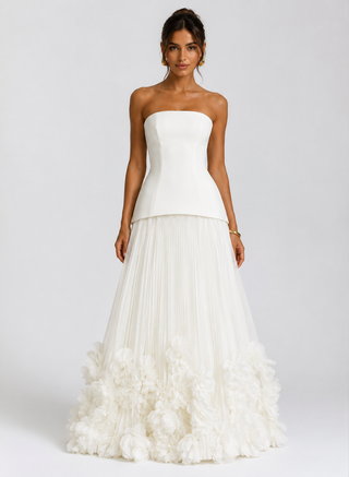 A woman modeling a strapless white gown with a structured corset-style bodice and a full pleated skirt featuring a ruffled floral border at the hem, photographed against a light gray studio background.