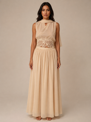 A woman modeling a sleeveless maxi dress in a soft champagne beige color, featuring a high neck with draped fabric, a lace-embellished waist, and a flowing skirt, set against a solid neutral studio background.