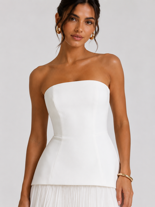 A woman wearing a strapless white corset-style top with a structured bodice and a pleated white skirt, photographed against a light gray studio background.