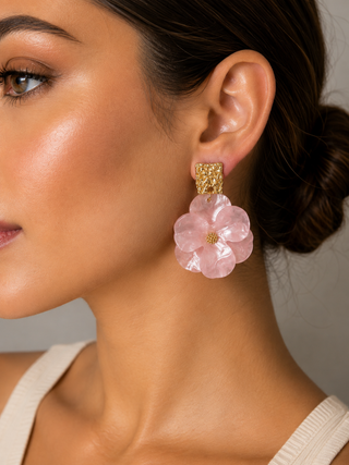 A close-up of a woman wearing a dangling earring with a textured gold top and a layered pink flower-shaped drop, shown against a neutral gray background.