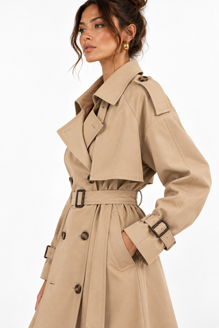 A woman wearing a classic beige trench coat with a matching waist belt and dark brown buttons, shown from a side profile against a plain white background.