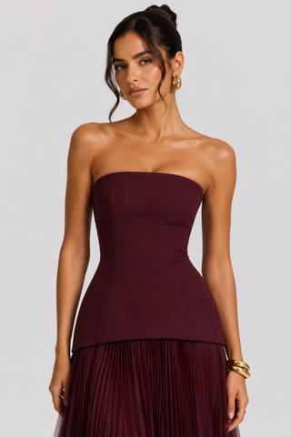 A woman modeling a strapless tube top and pleated skirt set in a deep burgundy or plum color, photographed against a light gray studio background.