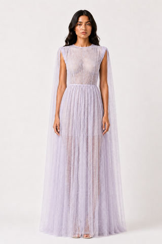 A model wearing a lavender lace and tulle maxi dress with a sheer cape-like overlay, photographed against a plain light gray studio background.