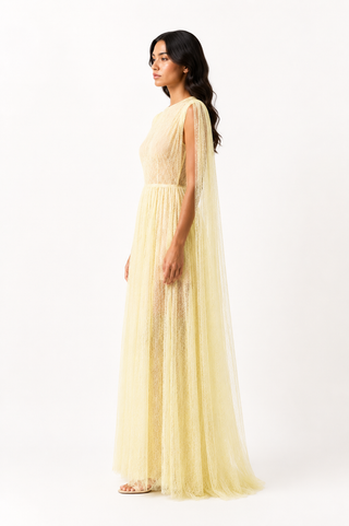 A woman modeling a floor-length pale yellow gown with a sheer lace bodice, draped cape sleeves, and a flowing tulle skirt against a plain white studio background.