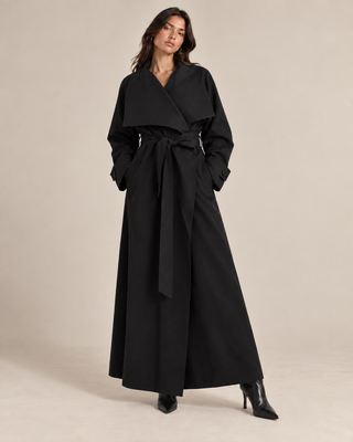 A woman modeling a long black wrap-style coat with a wide collar, long sleeves, a self-tie belt, and wide legs, paired with black pointed-toe heeled boots against a plain beige studio background.