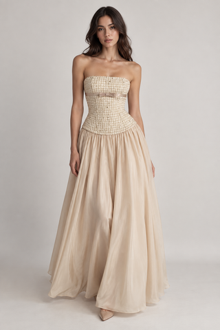 A woman modeling a strapless formal gown with a textured, woven-look bodice in beige and a flowing chiffon skirt in a light champagne beige, set against a plain light gray studio background.