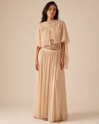 A woman modeling a two-piece beige maxi dress set featuring a sheer draped capelet overlay and a matching pleated maxi skirt, with a lace-trimmed crop top underneath, set against a solid warm beige studio background.