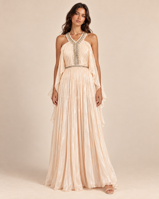 A woman modeling a floor-length halter gown in a warm cream or champagne tone with metallic embellished detailing at the neckline and waist, photographed against a plain beige studio background.