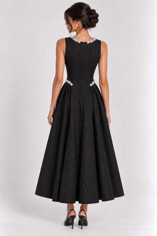 A woman modeling a sleeveless black A-line maxi dress from the back, featuring a beaded neckline and beaded waistband, paired with black heels against a light gray studio background.
