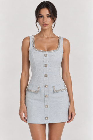 A woman modeling a light gray tweed mini dress with silver rhinestone trim on the neckline, pockets, and buttons, set against a plain light gray studio background.