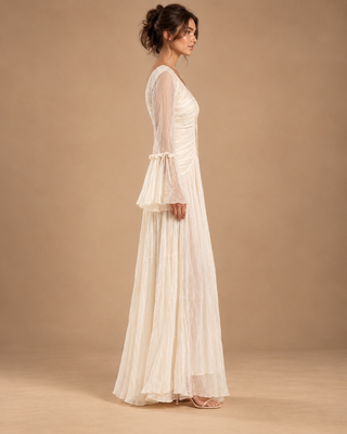A woman modeling a long white maxi dress with sheer long sleeves, ruffled detailing at the cuffs, and a flowing pleated skirt, shown against a solid beige studio backdrop.