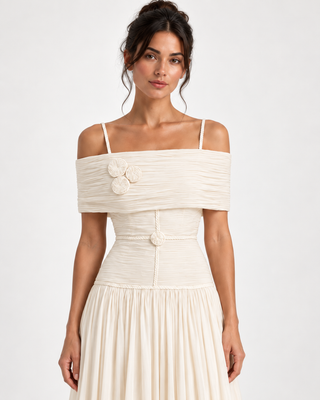 A woman modeling an off-the-shoulder cream-colored dress with a textured, gathered bodice, thin shoulder straps, and a pleated skirt against a plain light background.