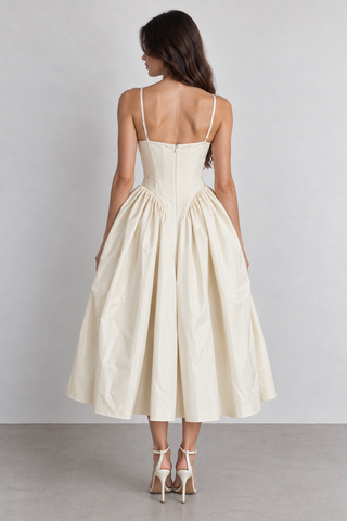 A woman shown from the back wearing a cream-colored midi dress with thin spaghetti straps, a structured corset-style back, and a full gathered skirt, paired with white high-heeled sandals against a neutral studio backdrop.