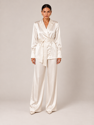 A model stands against a neutral beige studio background wearing a matching cream satin two-piece set consisting of a belted wrap jacket and wide-leg trousers.