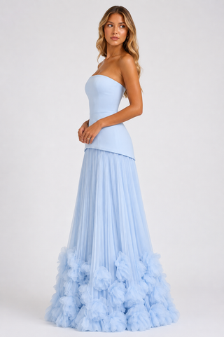 A woman modeling a strapless light blue gown with a fitted bodice and a voluminous pleated skirt featuring a ruffled tulle hem, photographed against a plain light gray studio background.