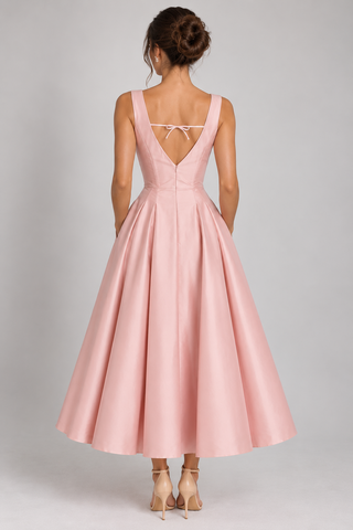 A woman shown from the back wearing a sleeveless pink A-line dress with a deep V back, tie detail at the neckline, and a full skirt, paired with nude high heels against a light gray studio background.