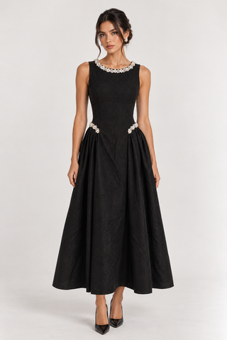 A woman modeling a sleeveless black midi dress with a beaded collar and matching beaded waistband, paired with black pointed heels against a light gray studio background.
