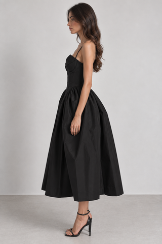A woman modeling a black midi dress with a structured strapless bodice and a voluminous gathered skirt, paired with black high-heeled sandals against a light gray studio background.