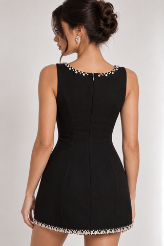 A woman shown from the back wearing a fitted black sleeveless mini dress with decorative pearl-like embellishments around the neckline and hem, against a plain light gray studio background.