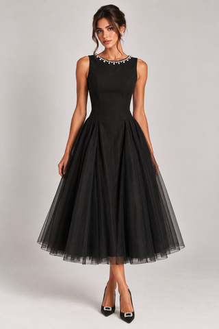 A woman modeling a sleeveless black midi dress with a fitted bodice, embellished neckline, and full tulle skirt, paired with black pointed heels against a light gray studio background.