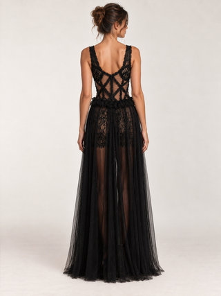 A woman modeling a black lace and tulle formal gown from the back, showing a crisscross open back bodice and a sheer layered skirt, against a plain light gray studio background.