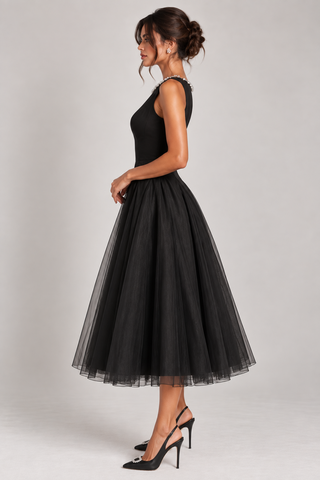 A woman modeling a sleeveless black midi dress with a fitted bodice, sheer embellished neckline, and a voluminous black tulle skirt, paired with black high heels against a light gray studio background.